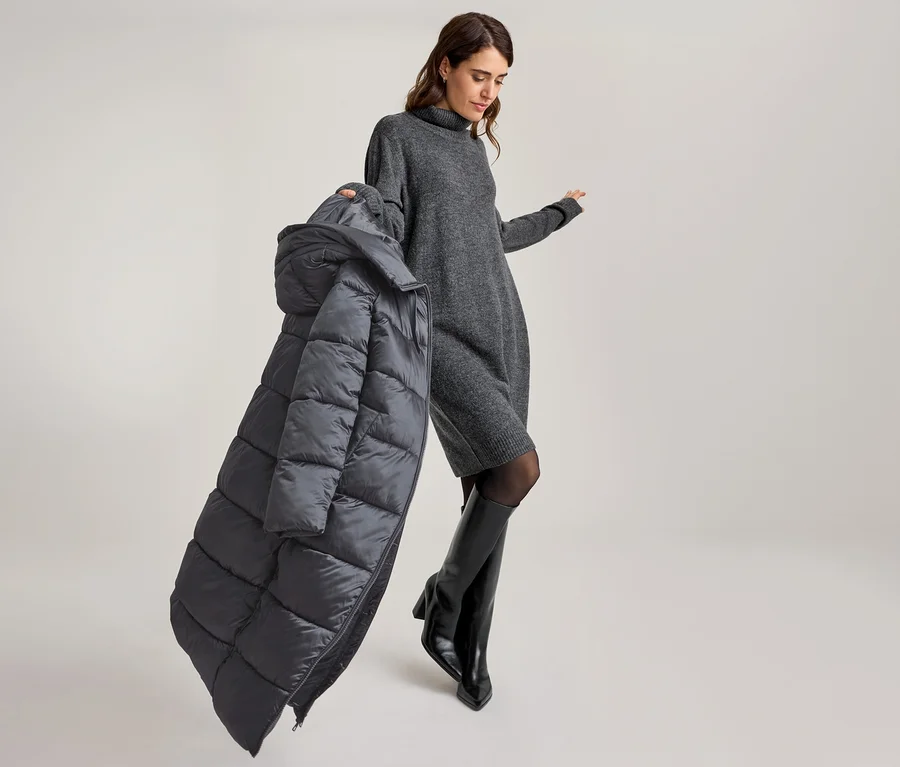 Femme en robe pull grise et bottes noires, tenant un manteau matelassé gris foncé.