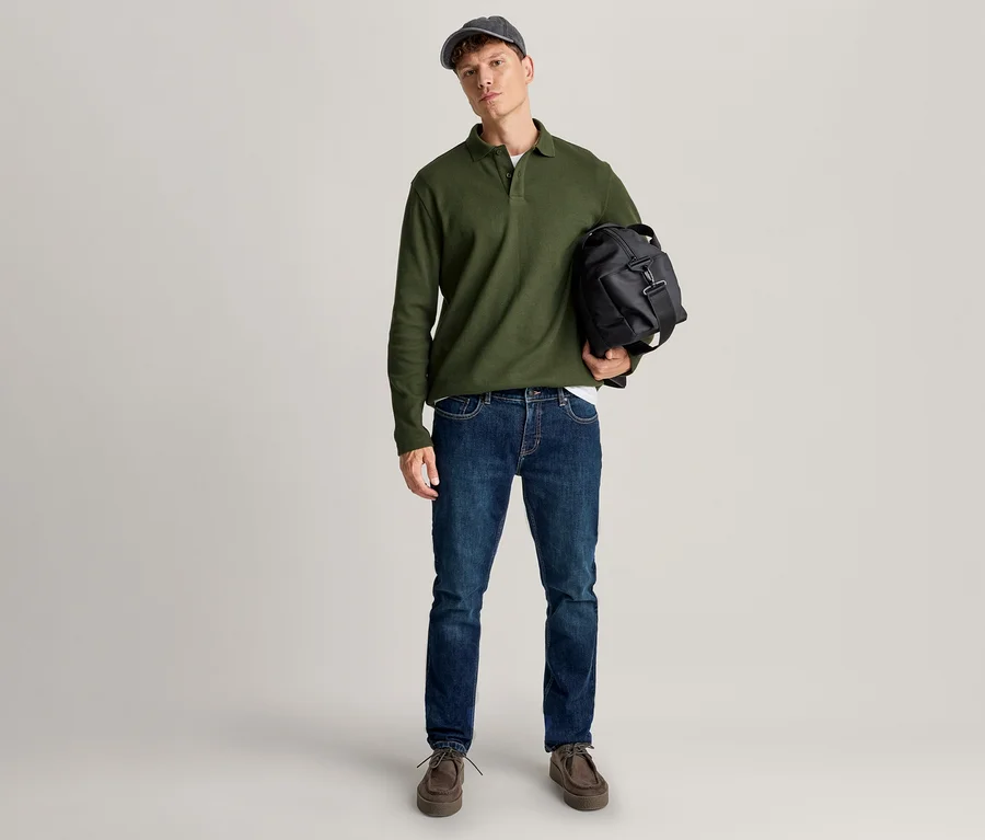 Un homme se tient debout, portant un polo vert à manches longues, un jean bleu, des chaussures marron et une casquette foncée, tenant un sac de voyage noir.