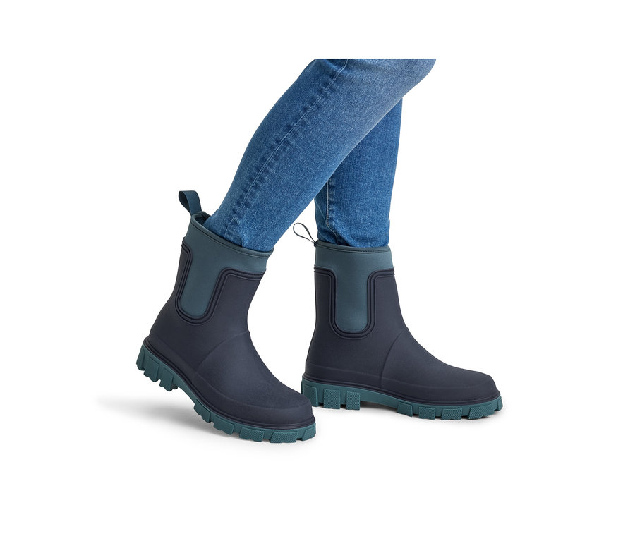 Personne portant des bottes de pluie bleu foncé avec des semelles sarcelle et des empiècements élastiques sarcelle aux chevilles, associées à un jean bleu.