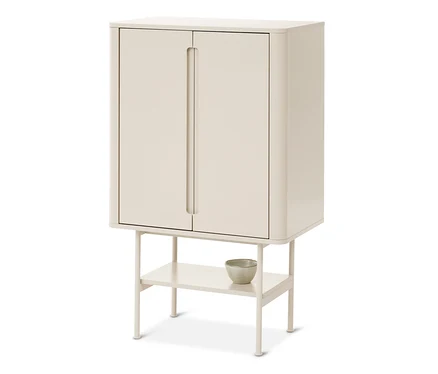 Une armoire couleur crème avec deux portes et des coins arrondis, reposant sur quatre pieds avec une étagère ouverte en dessous. Un petit bol de couleur claire est posé sur l'étagère.