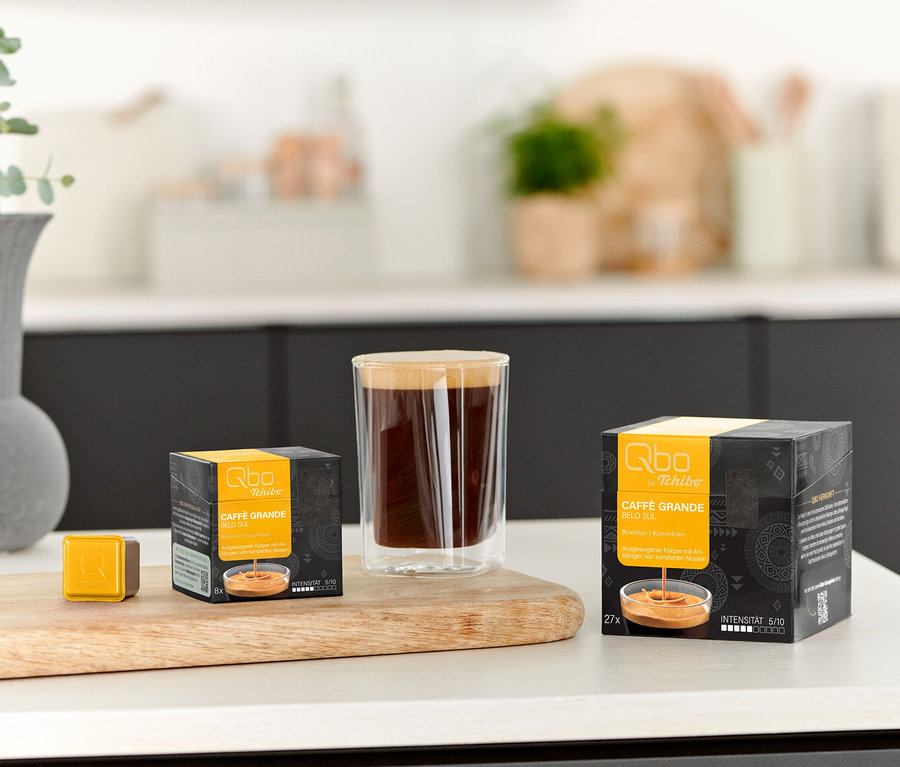Un verre de café est posé à côté d'un paquet de Qbo Caffè Grande BELO SUL - 8 capsules et d'une capsule individuelle sur une planche en bois.
