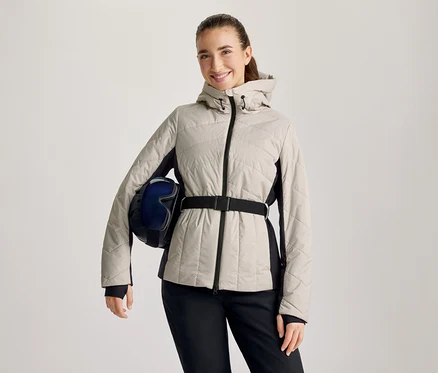 Femme souriante portant une veste matelassée beige avec des panneaux latéraux noirs et une ceinture noire, un pantalon noir, tenant un casque de ski bleu foncé avec des lunettes sous son bras gauche.