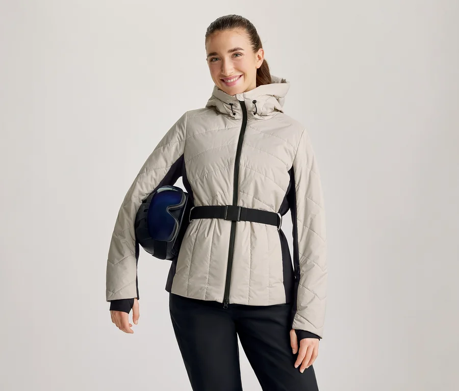 Femme souriante portant une veste matelassée beige avec des panneaux latéraux noirs et une ceinture noire, un pantalon noir, tenant un casque de ski bleu foncé avec des lunettes sous son bras gauche.