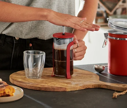 Femme souriante à lunettes pressant le piston d'une cafetière à piston rouge de 300 ml sur une planche en bois, avec des roulés à la cannelle et un verre vide.