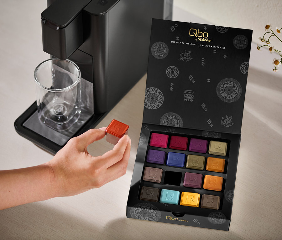 Une main tient une capsule de café rouge à côté d'une machine à café noire Qbo ICONIC Dark Chrome avec une tasse en verre et une boîte ouverte Qbo Collection - 16 capsules contenant diverses capsules colorées.