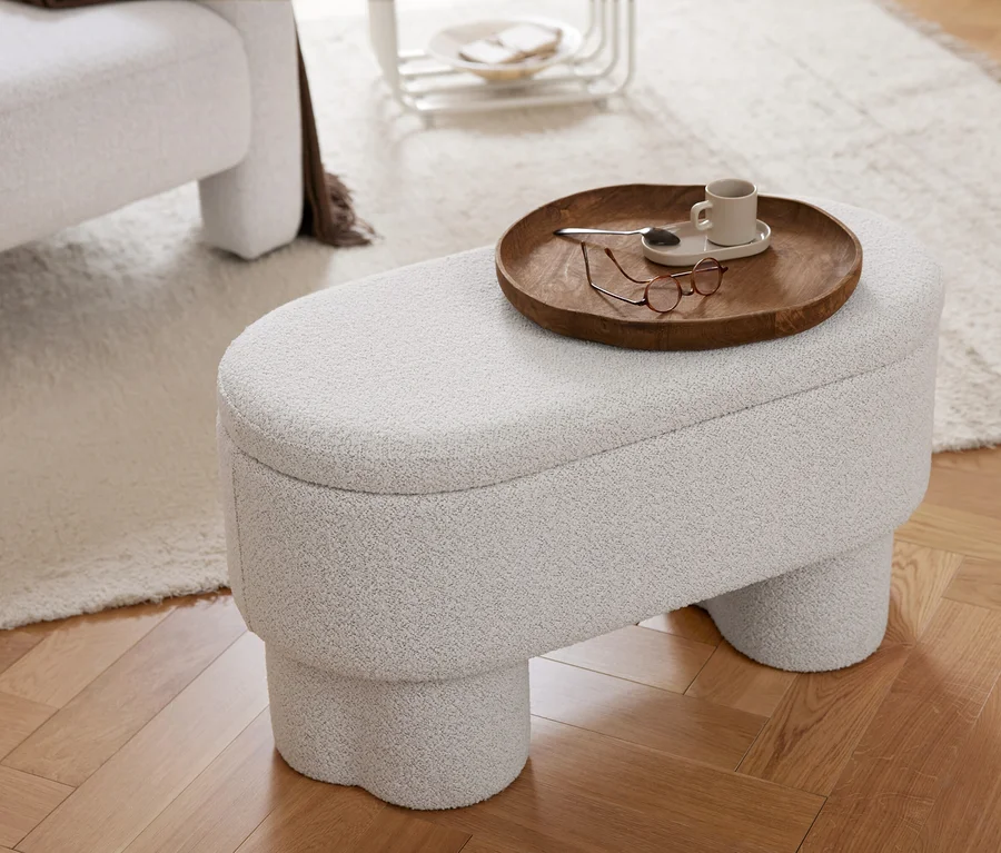 Un pouf texturé gris clair avec un plateau en bois contenant une tasse, une cuillère et des lunettes. À côté, un fauteuil avec un plaid marron, le tout sur un tapis clair et un parquet en chevrons.