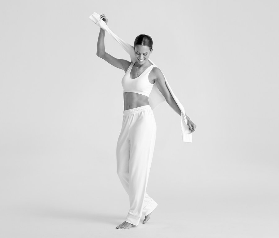 Femme souriante portant une brassière sans coutures crème et un pantalon, tenant un tissu au-dessus de sa tête.
