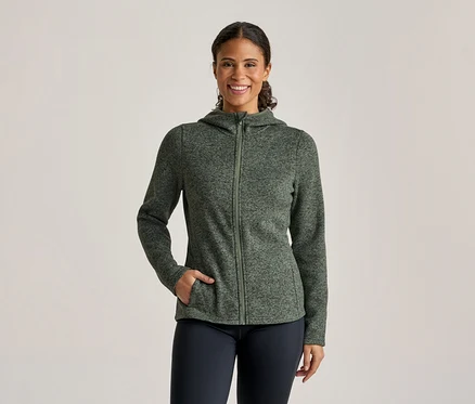 Une femme souriante porte une veste à capuche zippée verte mouchetée et un pantalon foncé, sa main gauche dans sa poche.