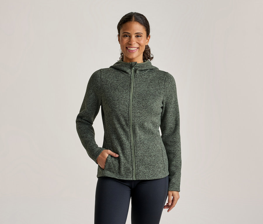 Une femme souriante porte une veste à capuche zippée verte mouchetée et un pantalon foncé, sa main gauche dans sa poche.
