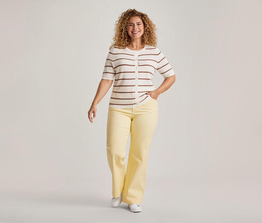 Une femme souriante aux cheveux bouclés avance, vêtue d'un cardigan crème à manches courtes avec des rayures marron, d'un pantalon large jaune et de chaussures blanches. Une main est dans sa poche.