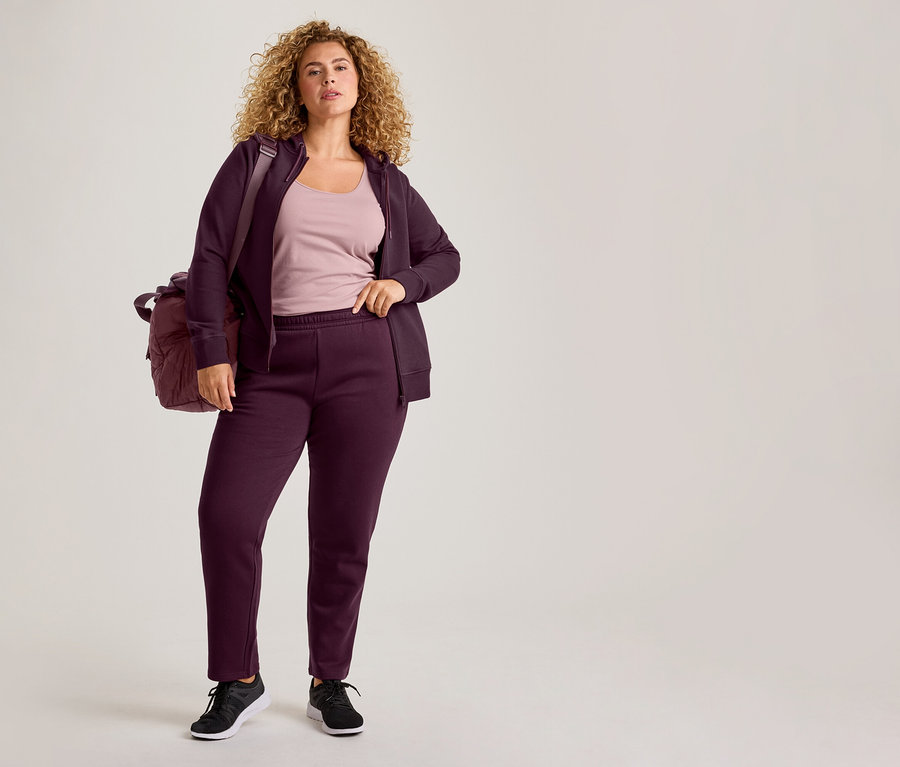 Une femme aux cheveux bouclés porte un sweat à capuche zippé violet, un pantalon de jogging assorti, un débardeur rose clair et des baskets noires, avec un sac de sport violet sur l'épaule.