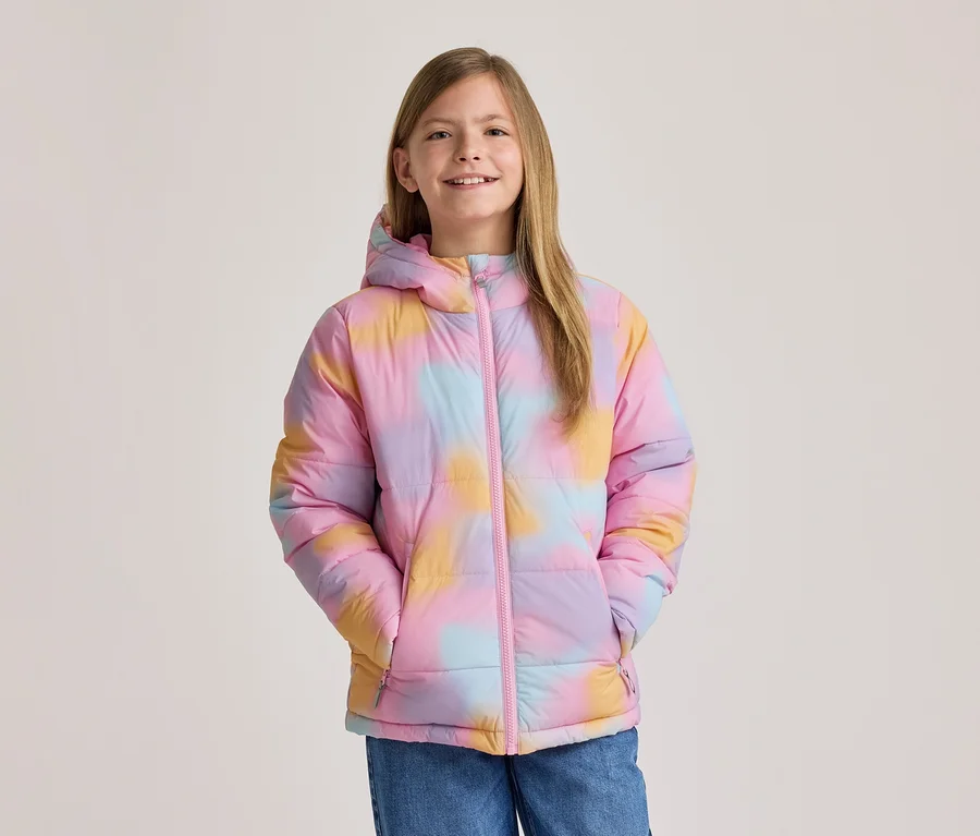Jeune fille souriante portant une doudoune tie-dye colorée rose, jaune et bleue, avec une capuche et une fermeture éclair rose, mains dans les poches.