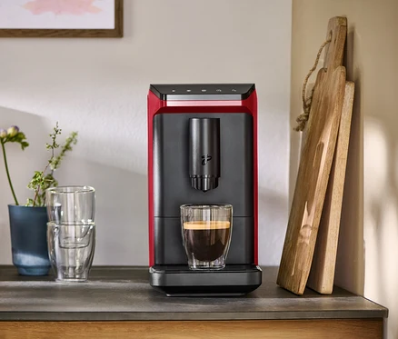 Machine à café entièrement automatique « Esperto mini », Lovely Red, rouge et noire, avec une tasse de café sur un comptoir, à côté de verres et de planches à découper.