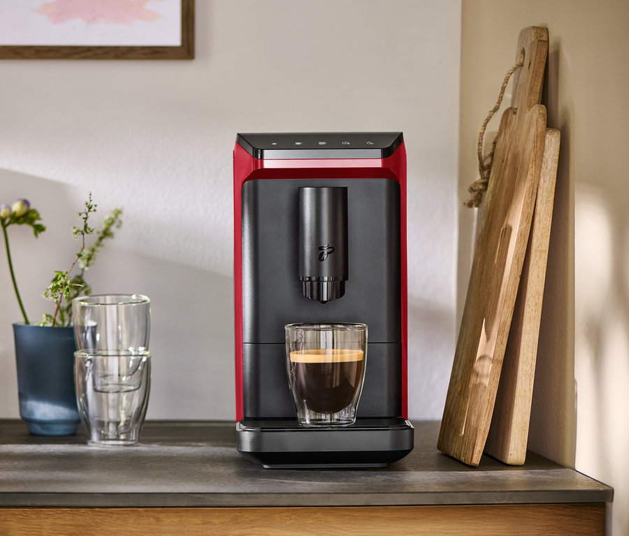 Machine à café entièrement automatique « Esperto mini », Lovely Red, rouge et noire, avec une tasse de café sur un comptoir, à côté de verres et de planches à découper.