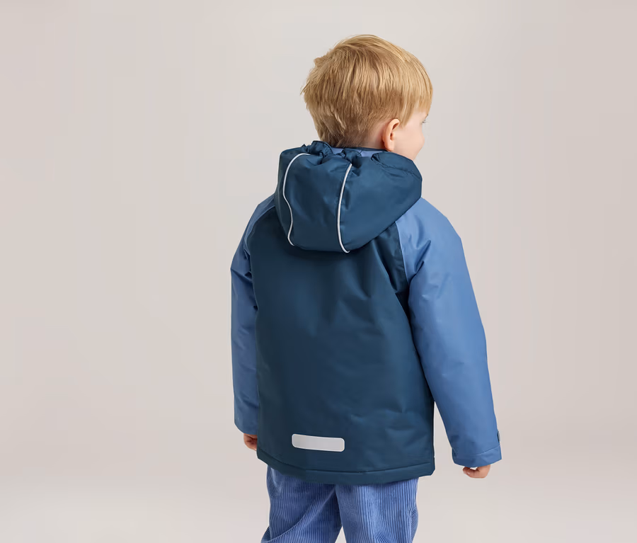 Jeune enfant vu de dos, portant une veste à capuche bicolore bleue avec des détails réfléchissants et un pantalon en velours côtelé bleu clair. L'enfant a les cheveux blonds et regarde vers la droite.