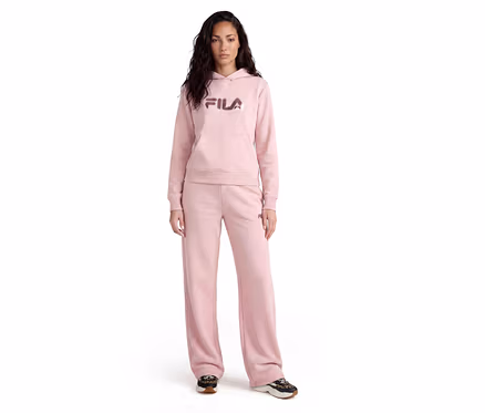 Femme debout, portant un sweat à capuche Fila rose avec une poche kangourou et un pantalon de jogging large assorti, associé à des baskets foncées.