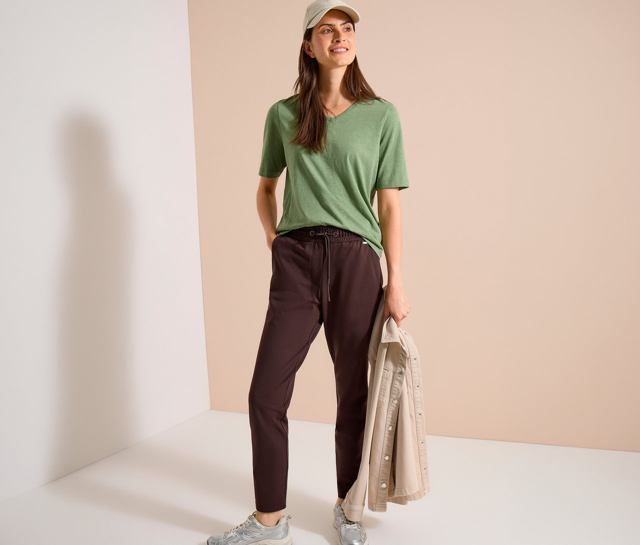 Une femme avec une casquette beige porte un t-shirt vert à col en V, un pantalon en jersey CECIL marron et des baskets argentées, tenant une veste beige.