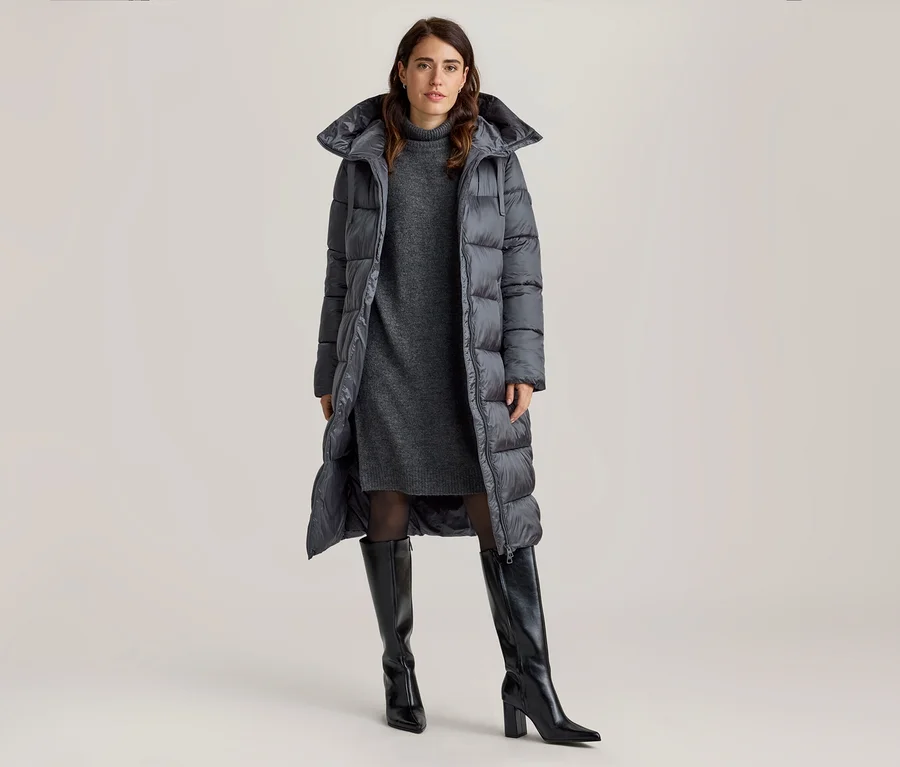 Femme portant un manteau matelassé extra-long sur une robe pull grise et des bottes noires montantes.