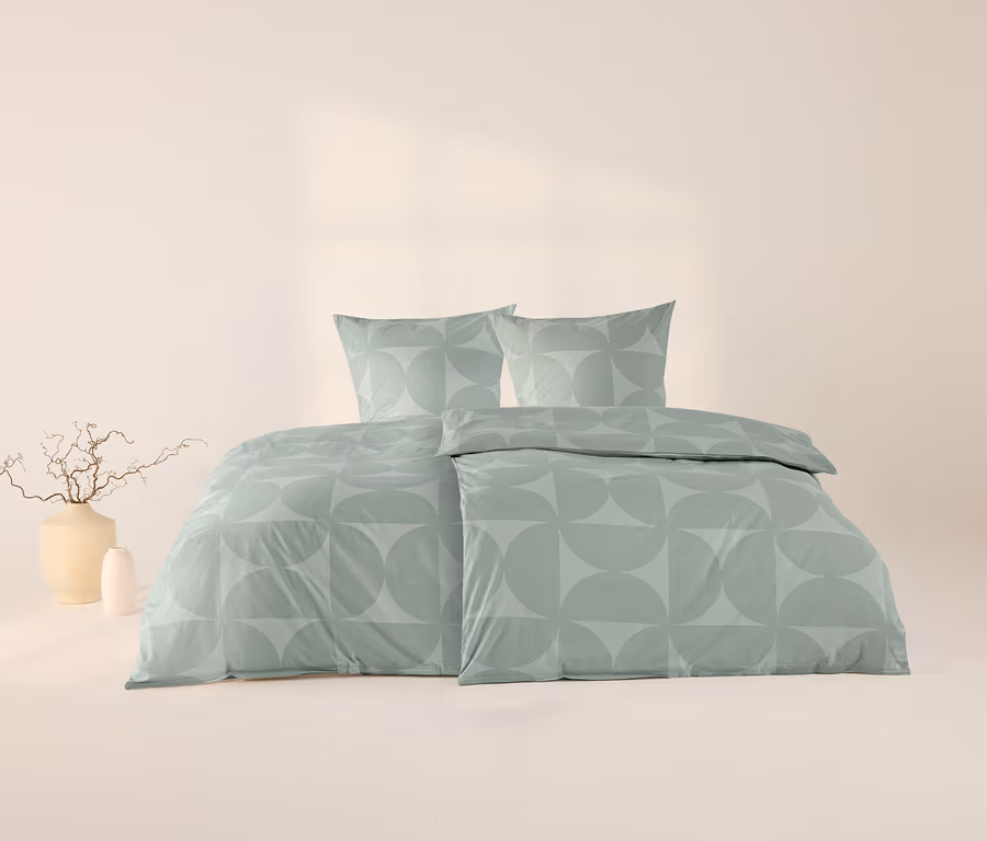 Parure de lit en renforcé verte avec un motif géométrique de cercles et demi-cercles, comprenant deux oreillers et une housse de couette, sur fond clair avec deux vases décoratifs à gauche.