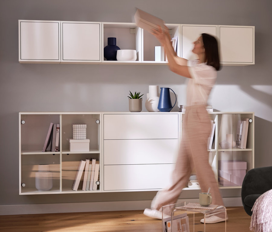 Une femme place une boîte dans une étagère murale. Au premier plan, une table d'appoint avec porte-revues et des étagères modulables « Flemming ».