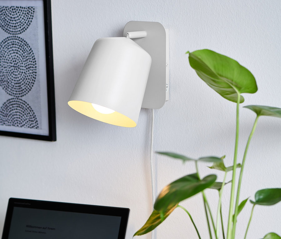 Une lampe murale en métal blanche éclaire un bureau avec un ordinateur portable et une plante.