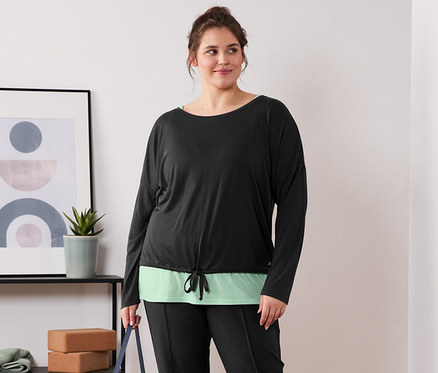 Une femme debout, pieds nus, tenant un tapis de yoga. Elle porte un t-shirt de sport 2 en 1 noir et vert et un pantalon de jogging noir.