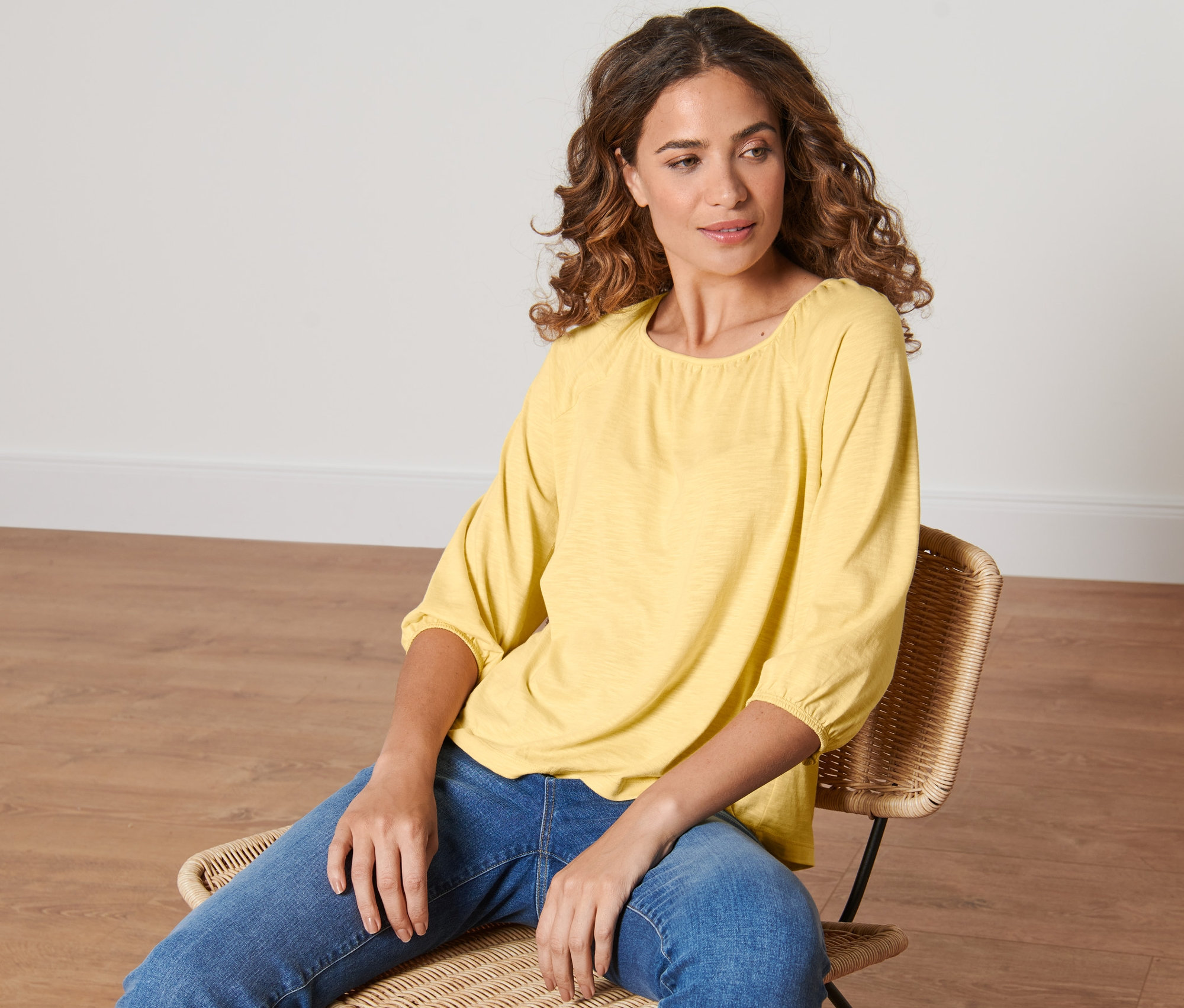 Une femme est assise sur une chaise en rotin et porte une blouse jaune à manches 3/4.