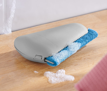 Une raclette pour angles et joints repose sur une surface en bois à côté d'un bol blanc et d'un chiffon rose.