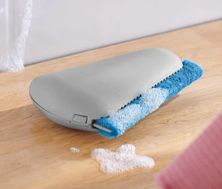 Une raclette pour angles et joints repose sur une surface en bois à côté d'un bol blanc et d'un chiffon rose.