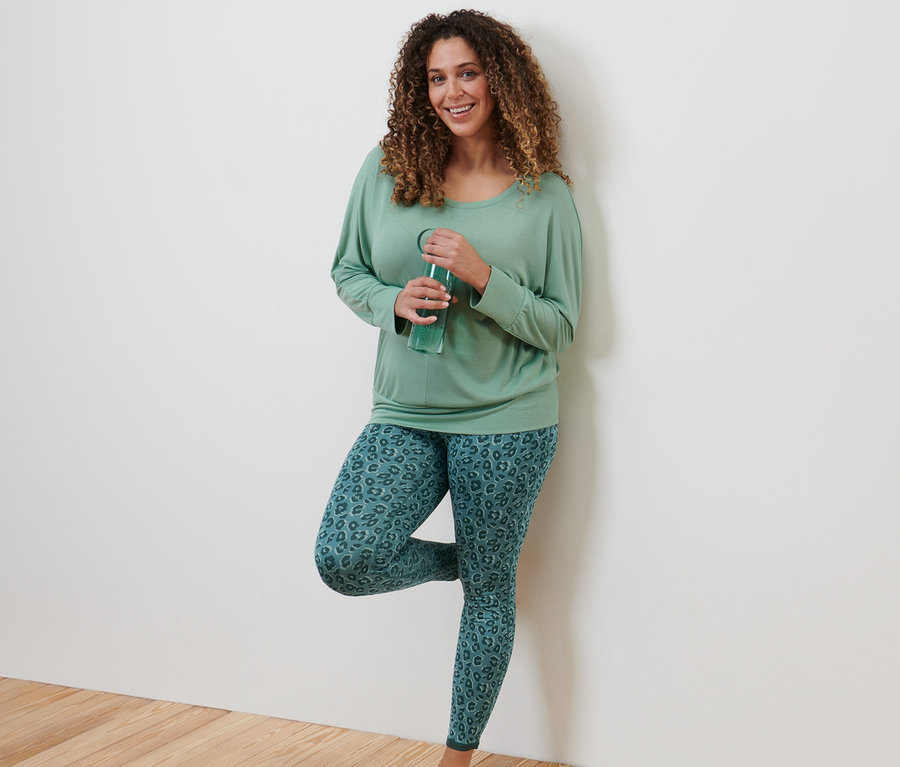 Une femme est appuyée contre un mur et boit de l'eau. Elle porte un T-shirt de sport et de yoga, vert sauge et un legging de sport réversible, émeraude et sauge.