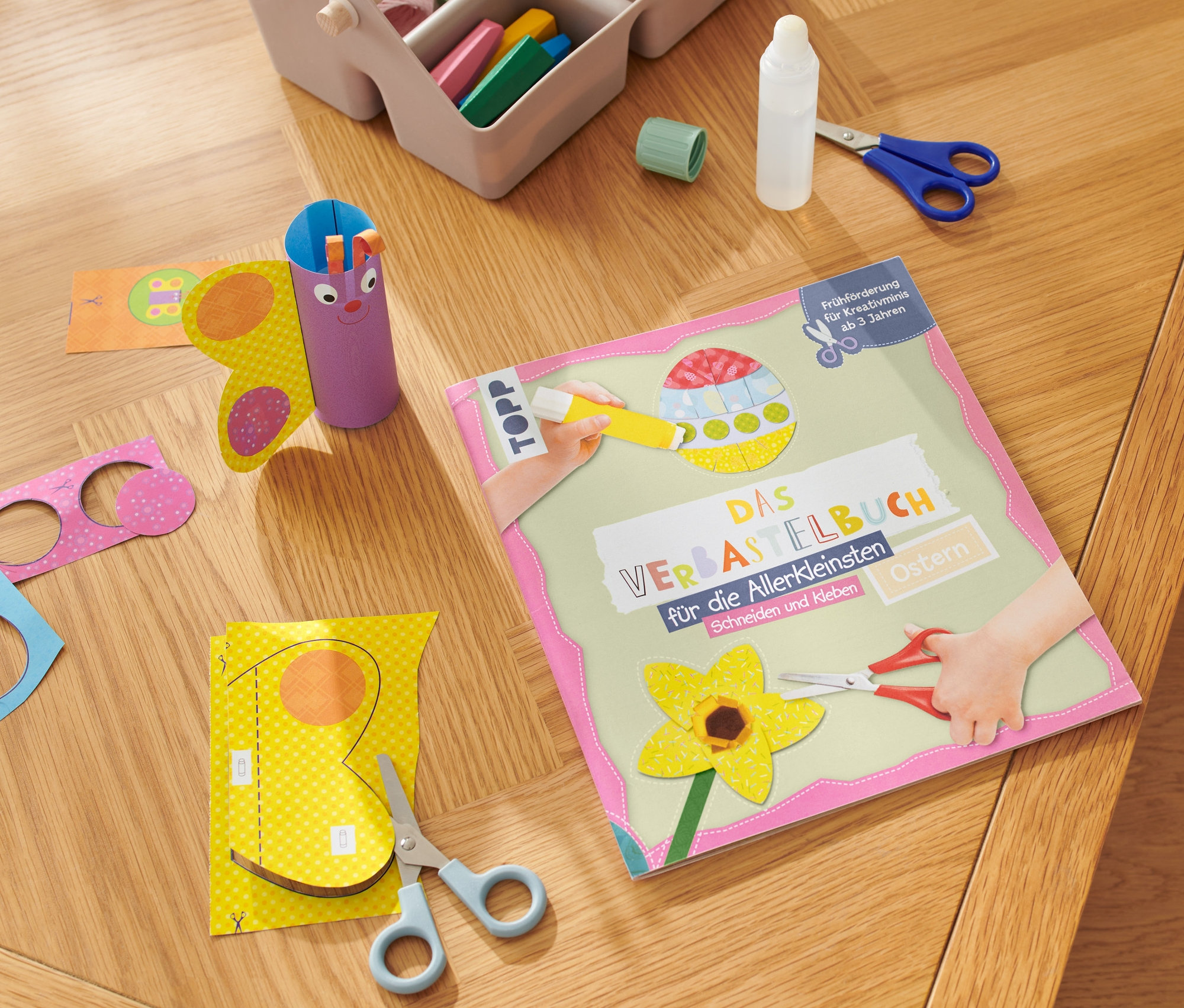 Sur une table en bois sont posés le livre « Das Verbastelbuch für die Allerkleinsten – Ostern », des ciseaux, de la colle et des travaux manuels en papier en cours de réalisation.