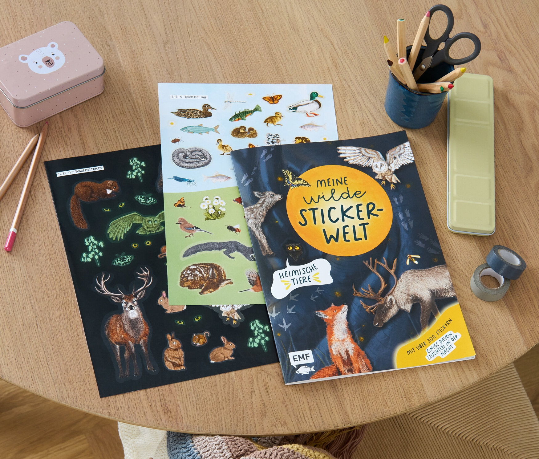 Sur une table en bois se trouvent des crayons, des ciseaux, une boîte et le livre d'autocollants «
Meine wilde Stickerwelt – heimische Tiere
».