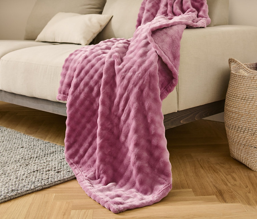 Une couverture moelleuse d'environ 150 x 200 cm est posée sur un canapé beige. Un panier en osier est placé à côté du canapé.