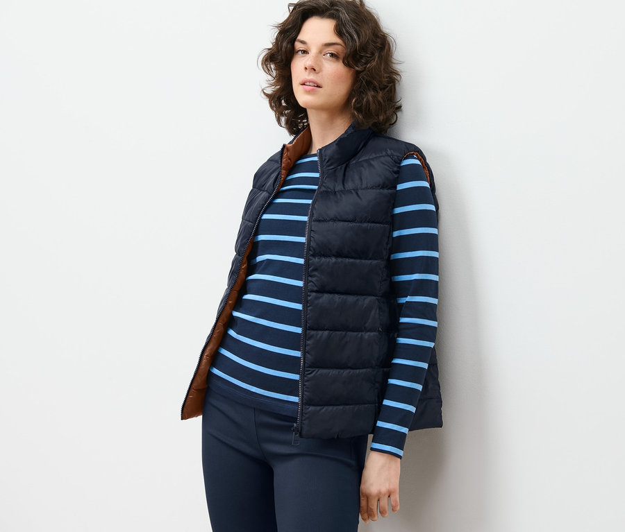 Femme appuyée contre un mur portant un gilet matelassé réversible, un t-shirt rayé et un pantalon.