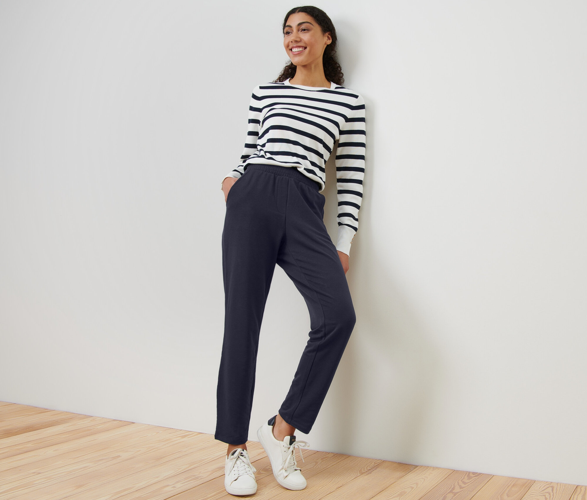 Une femme portant un pantalon de jogging bleu marine et un haut rayé est appuyée contre un mur.