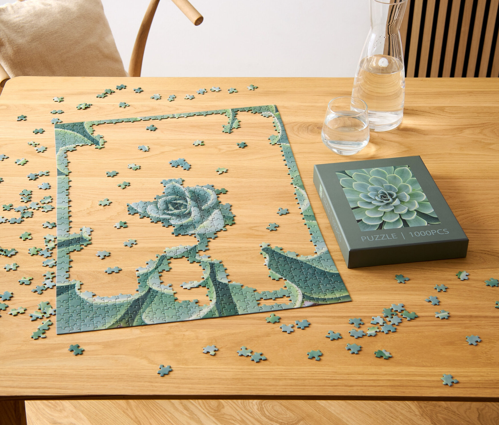 Un puzzle partiellement assemblé « Succulentes » est posé sur une table en bois à côté d'une boîte avec un motif de succulentes, d'un verre et d'une carafe d'eau.