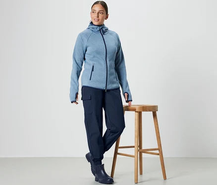 Une femme se tient à côté d'un tabouret en bois et porte un sweat à capuche bleu et un pantalon de jogging softshell.