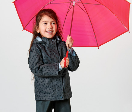 Une fille tient un parapluie rouge et porte une veste de pluie thermique pour tout petit, bleu ardoise gouttes, et un pantalon de pluie.