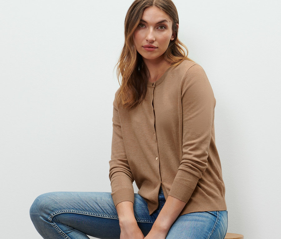 Une femme est assise sur un tabouret en bois et porte un cardigan en maille fine, un jean et des baskets.