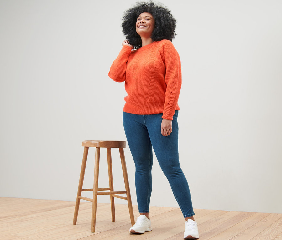 Une femme pose à côté d'un tabouret et porte un pull en maille orange et un jegging.