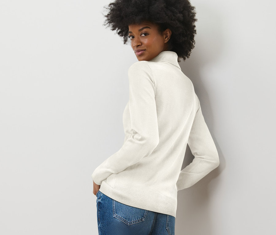 Femme regardant par-dessus son épaule portant un pull à col roulé en maille fine blanc crème et un jean.