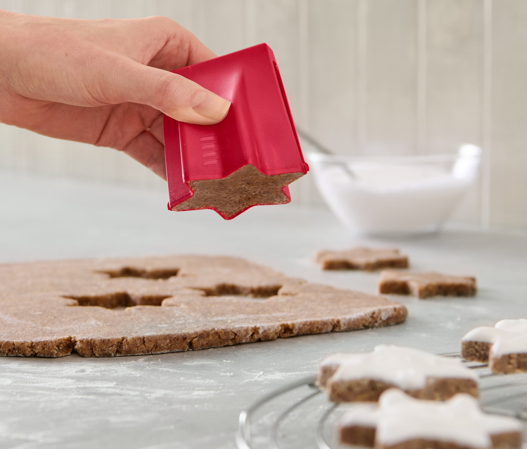 Une main utilise un emporte-pièce étoile à la cannelle sur de la pâte. Sur le comptoir, il y a des étoiles décorées et un bol de glaçage.
