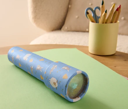 Un caléidoscope posé sur une feuille de papier verte à côté d'un pot contenant des crayons de couleur et des ciseaux.