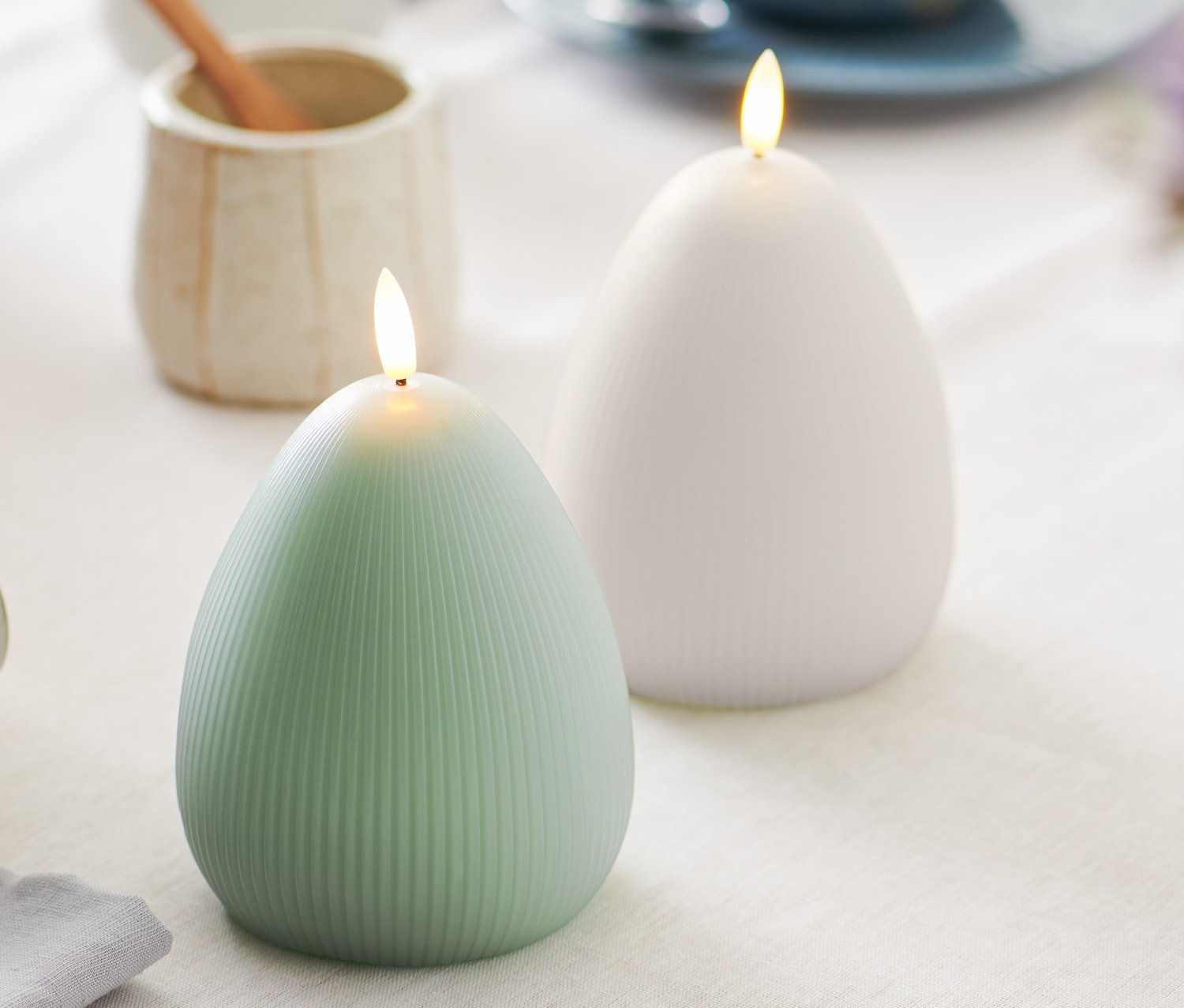 Sur une table, deux bougies à LED en cire véritable en forme d'œuf, une verte et une blanche.