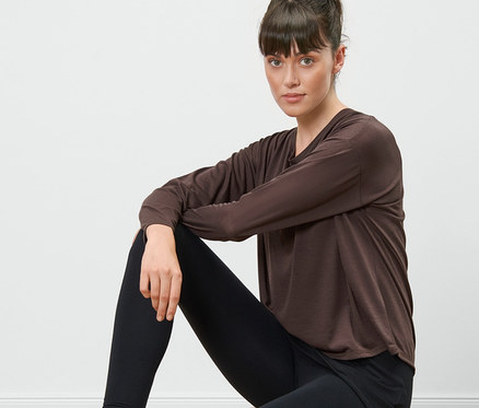 Une femme est assise sur un tabouret en bois et porte un t-shirt de sport 2 en 1 marron et un legging noir.