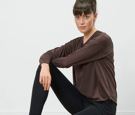 Une femme est assise sur un tabouret en bois et porte un t-shirt de sport 2 en 1 marron et un legging noir.