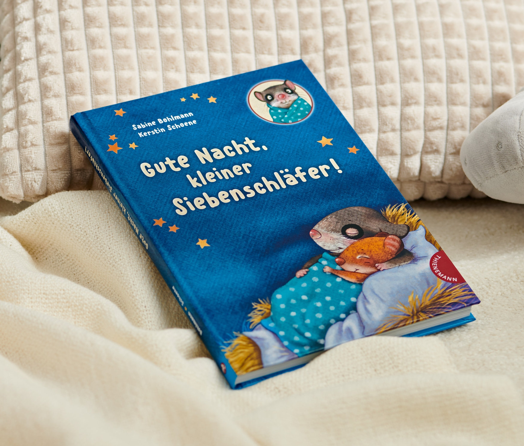 Le livre « Gute Nacht, kleiner Siebenschläfer ! » est posé sur une couverture à côté de coussins et d'un hippopotame en peluche.