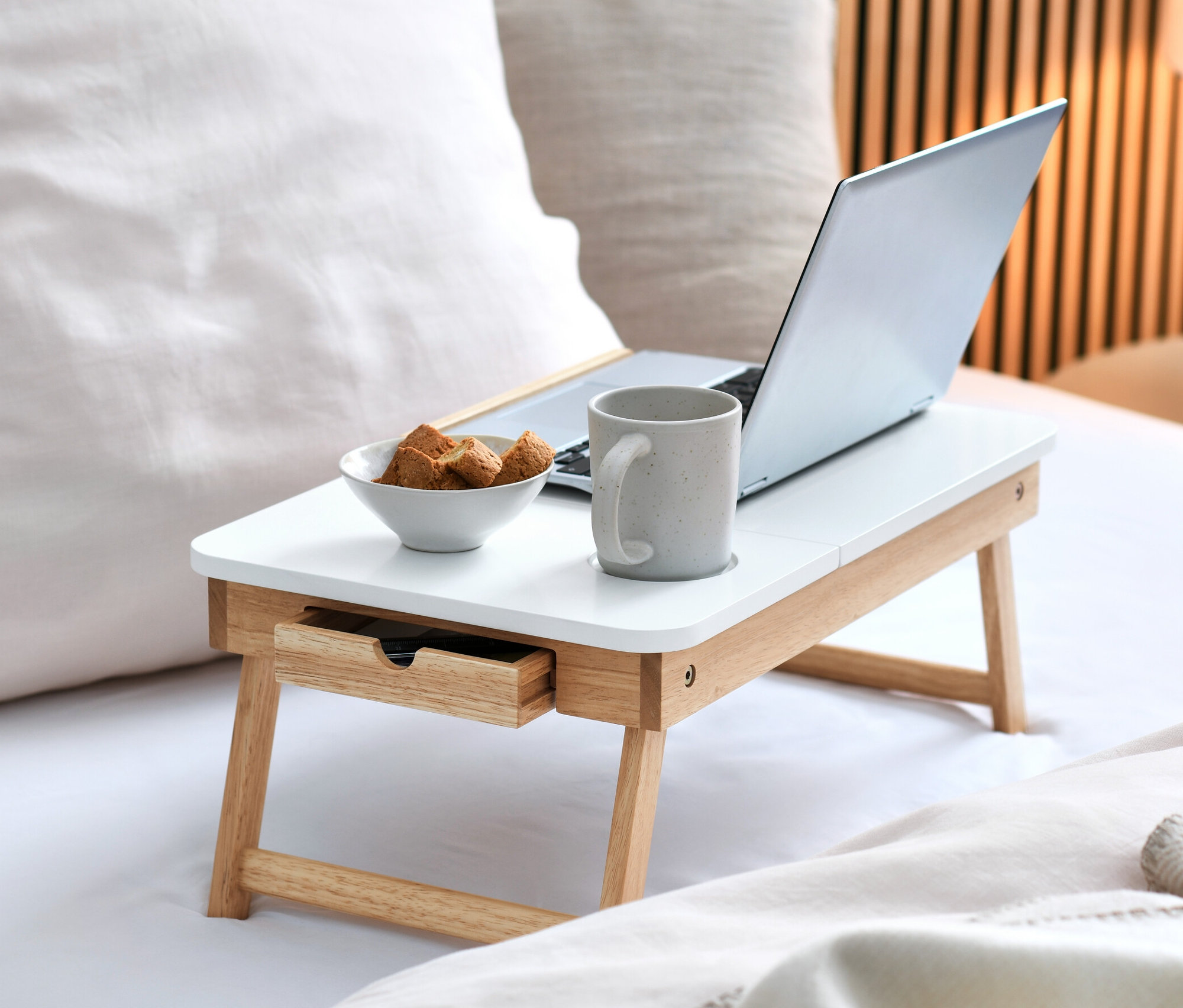 Sur un lit, une table de lit pour ordinateur portable avec un ordinateur portable, un bol de biscuits et une tasse.
