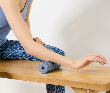 Une femme est assise sur un banc et touche avec une main un mini-rouleau pour fascias.