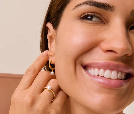 Une femme souriante touche une boucle d'oreille et porte une bague en argent 925, des créoles en argent 925 avec une perle de culture et une chaîne en or 375 billes.